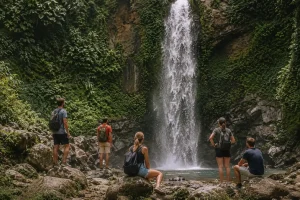 Tourists enjoying the natural beauty of Carat Waterfall in Bali, standing near the powerful cascading falls surrounded by lush tropical rainforest, misty air, and dramatic cliffside scenery while taking photos and relaxing in a pristine jungle setting.
