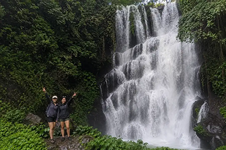 Two female hikers gesturing peace signs in front of a wide, multi-tiered waterfall cascading down a cliff.