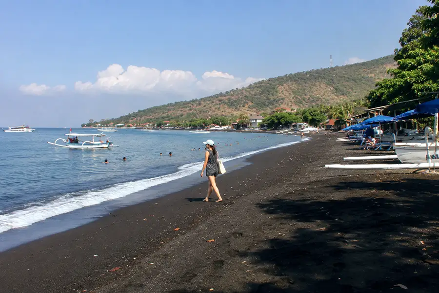 A tourist strolling along a black sand shoreline dotted with traditional boats and umbrella loungers.