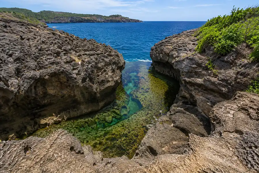 A clear, emerald-green tidal pool nestled between rugged volcanic rock cliffs leading out to the deep blue sea.