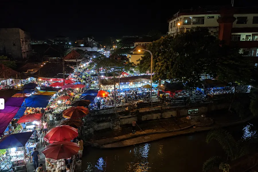 High-angle view of vibrant outdoor market stalls with colorful umbrellas along a river at night.