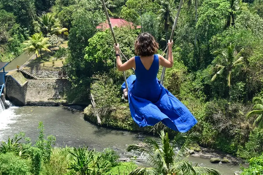 A woman in a flowing blue dress seen from behind swinging high over a tropical river valley.