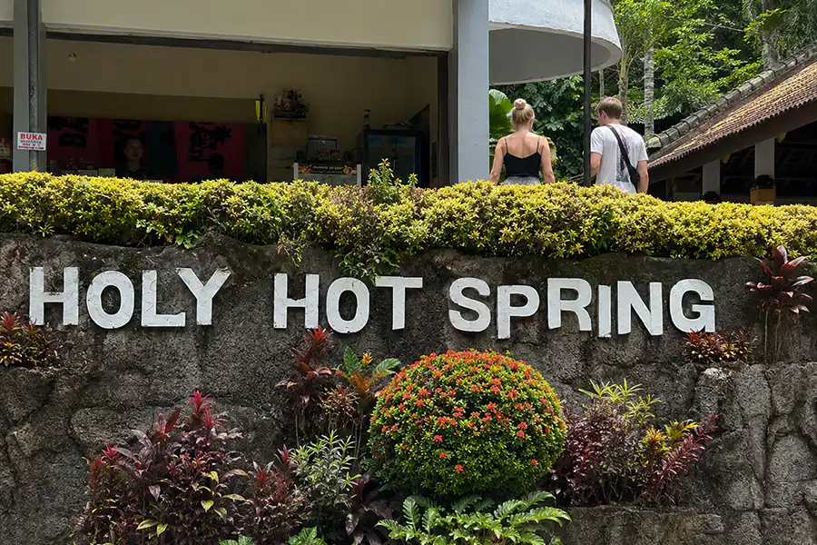 Tourists walking near a large stone wall reading "HOLY HOT SPRING" decorated with red and green plants.