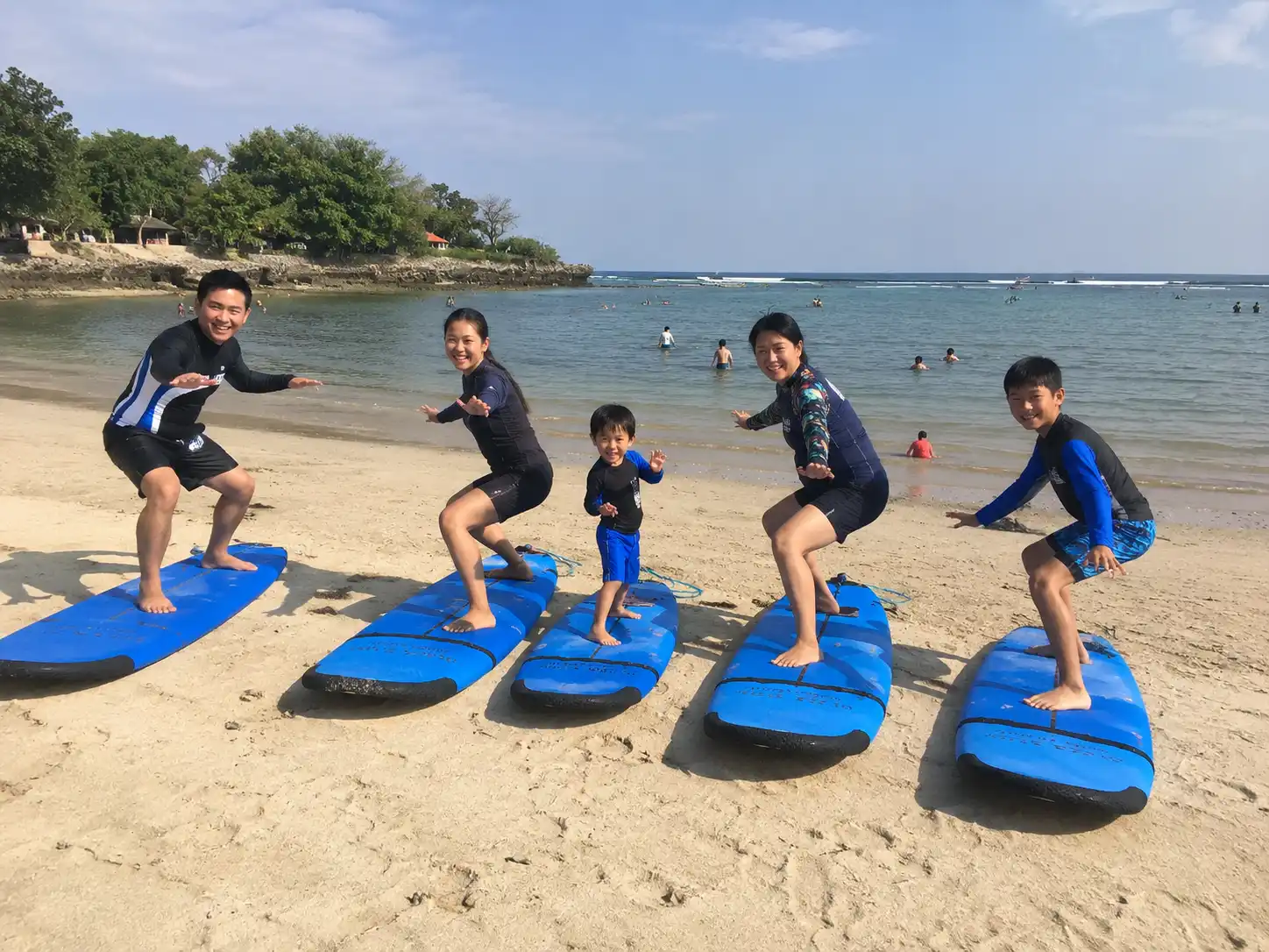 A family of five, including three children, pose in surfing stances on blue foam surfboards on a sunny beach in a sandy lagoon.