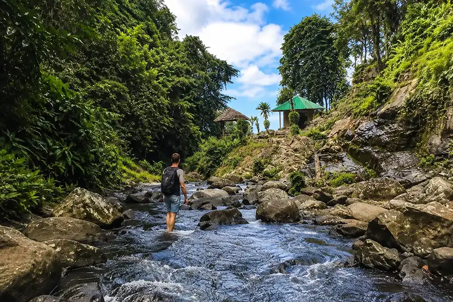 A male hiker with a backpack wading through a rocky shallow river toward wooden huts on a hill.