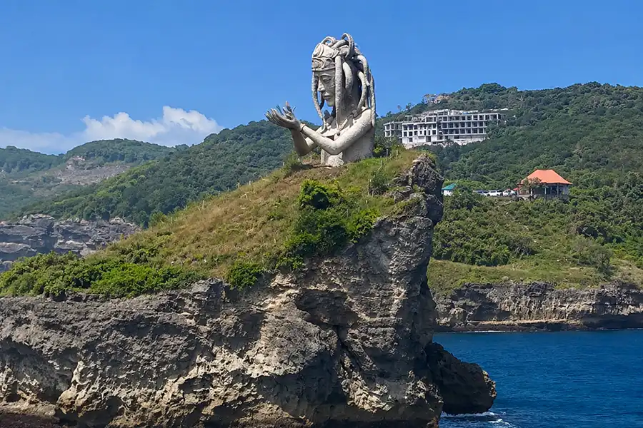 A giant stone statue of a figure with open hands sitting atop a grassy cliff by the ocean.