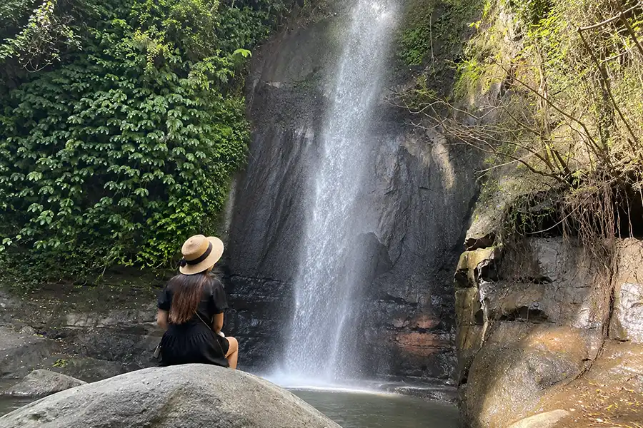 A woman wearing a straw hat and black dress sitting on a large boulder, looking up at a tall splashing waterfall.
