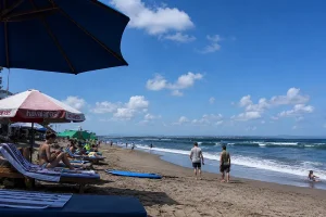 People relaxing on lounge chairs with beach umbrellas on a dark sand shoreline under a bright blue sky.