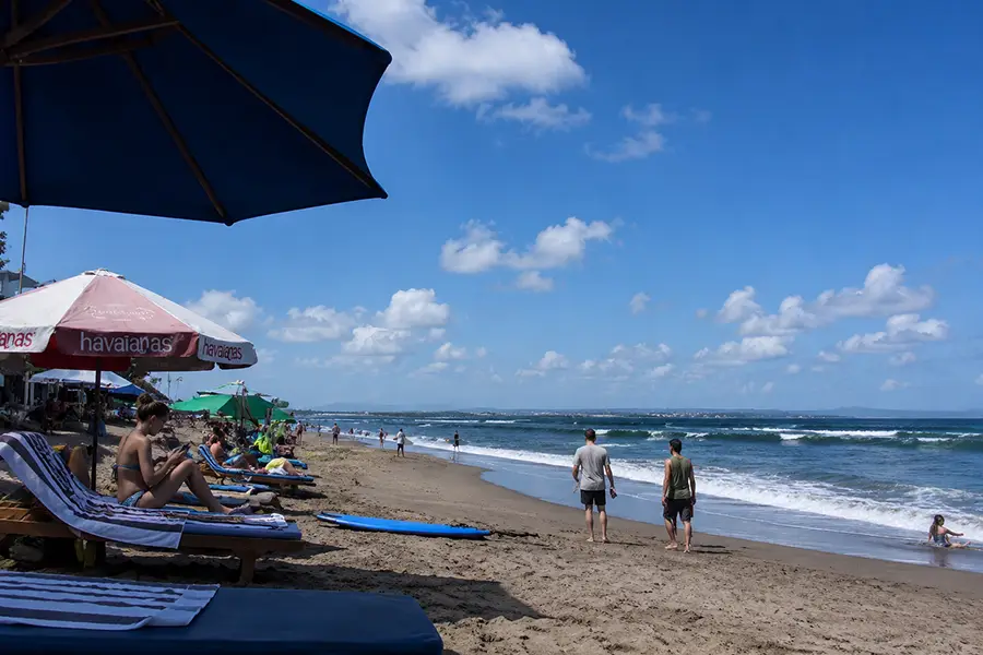 People relaxing on lounge chairs with beach umbrellas on a dark sand shoreline under a bright blue sky.