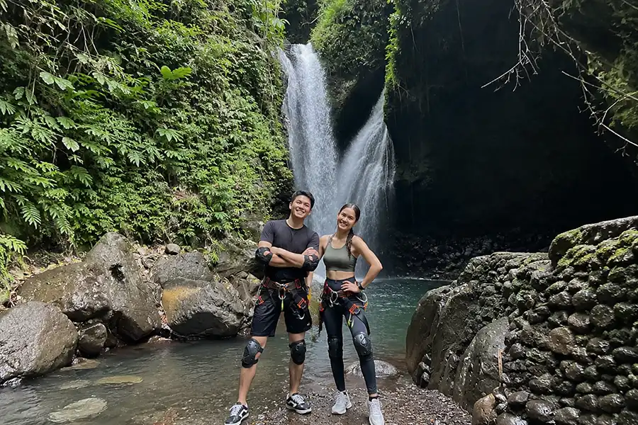 A man and woman wearing harness gear and knee pads posing on riverbank in front of a twin waterfall.