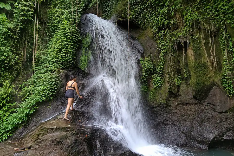 A woman standing on dark slippery rocks right next to a gushing upper waterfall stream.