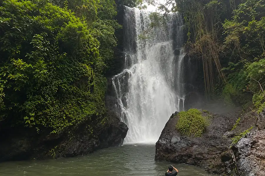 A photographer taking a picture of a wide, gushing waterfall dropping into a river pool surrounded by rocky cliffs and jungle vines.