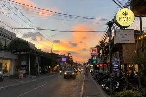 A Canggu street scene at sunset with cafe signs, parked scooters, and vehicles under a warm sky.