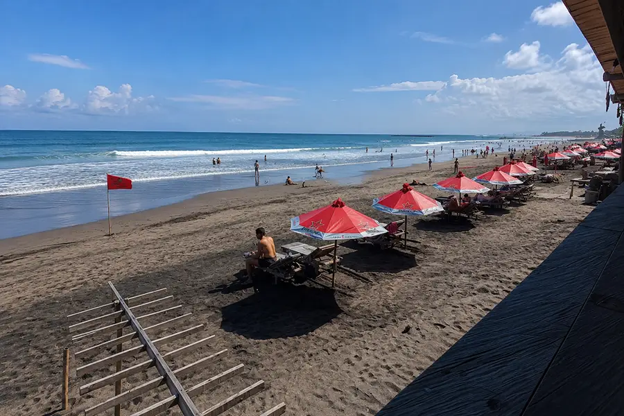 Tourists lounging on sunbeds under red umbrellas along a dark sand beach with ocean waves.