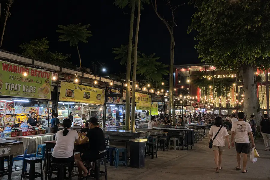 Visitors sitting at outdoor dining tables under warm fairy lights surrounded by local food stalls.
