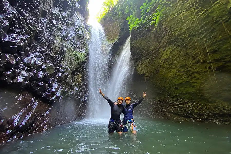 Two adventurers in helmets standing in a gorge pool with hands raised in front of a waterfall.