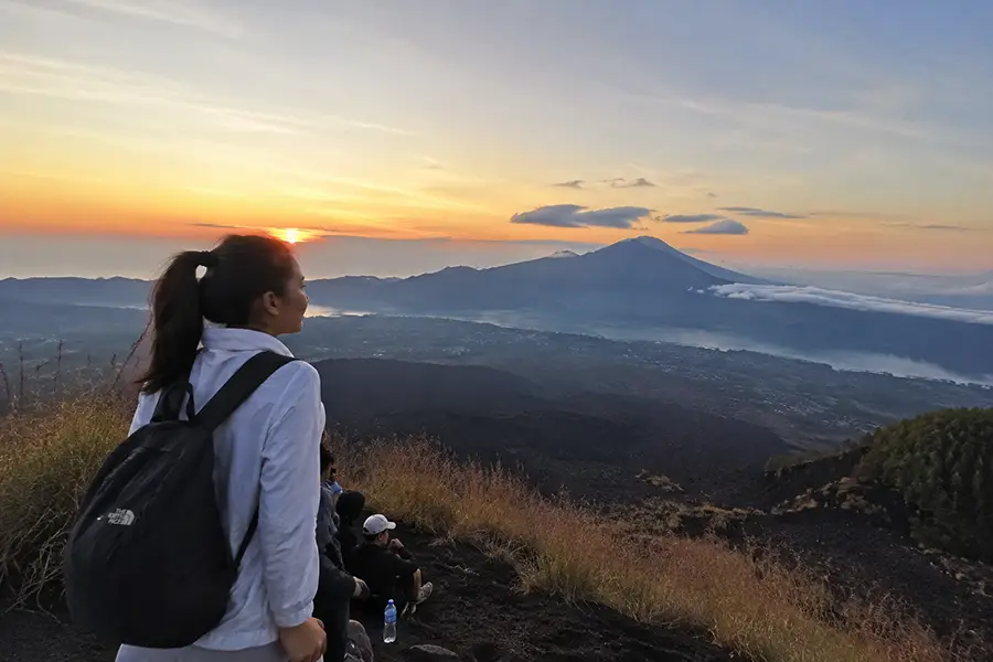 A woman with a backpack looking out at a volcanic caldera valley during sunrise.