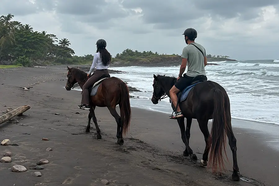 Two people wearing helmets riding horses along a black sand beach near ocean waves.