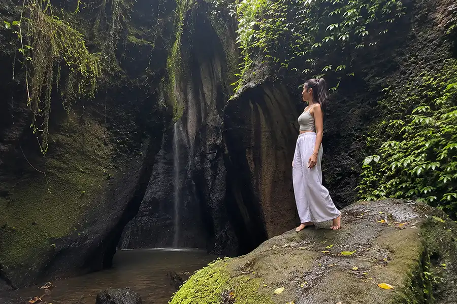 A woman in white pants standing on a mossy boulder inside a rocky canyon facing a hidden waterfall.