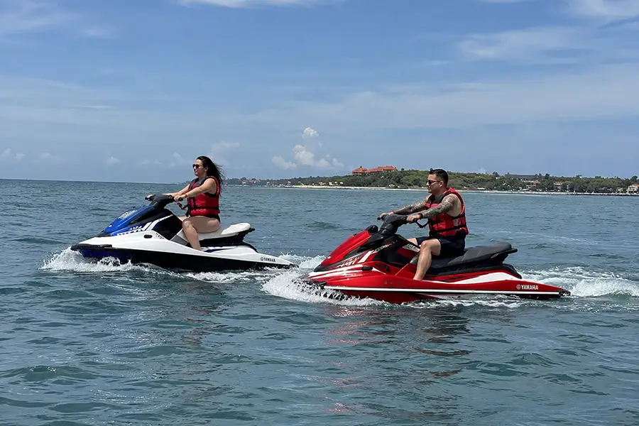 A man and a woman riding red and blue jet skis across open ocean waters under a sunny sky.