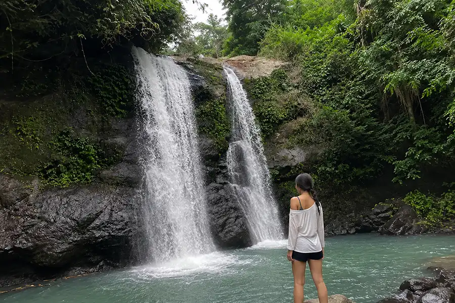 A woman standing near the water looking at twin waterfalls flowing down dark rock walls into a turquoise pool.