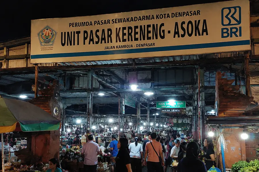 Shoppers walking past the entrance sign of Unit Pasar Kereneng Asoka surrounded by covered market stalls.