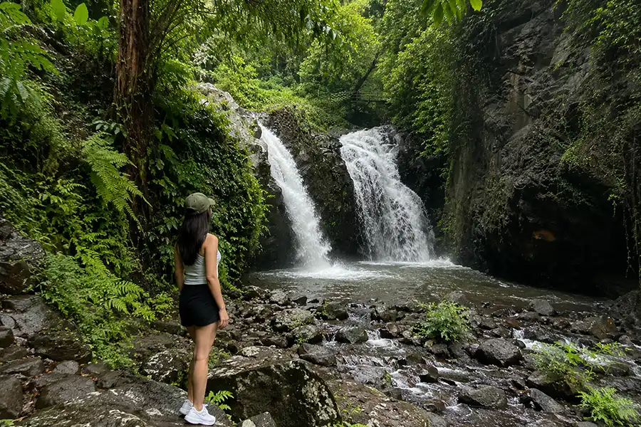 A woman standing on mossy river rocks looking toward a twin-stream waterfall flowing into a pool.