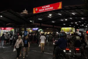 Visitors walking beneath a bright red "Kuliner Pasar Badung" sign surrounded by food stalls and motorbikes at night.