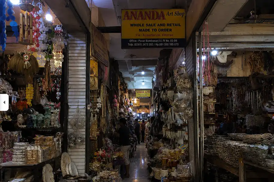 A narrow indoor market aisle filled with traditional wooden crafts, shell ornaments, and art shop signs.