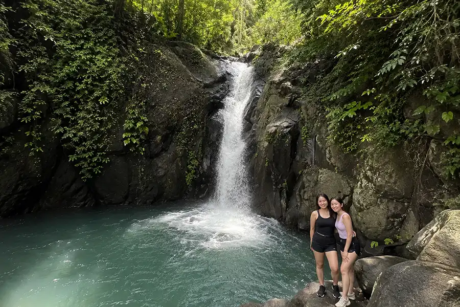Two women posing on rocks in front of a powerful waterfall dropping into a clear blue basin.