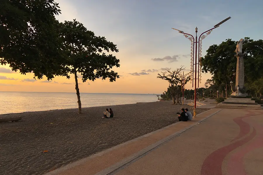 People sitting on the paved promenade and black sand beach next to the dolphin monument during sunset.