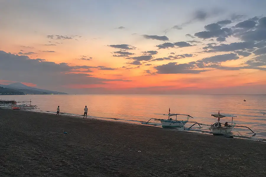 Traditional wooden jukung outrigger boats resting on the black sand shore under a vibrant orange sunset sky.