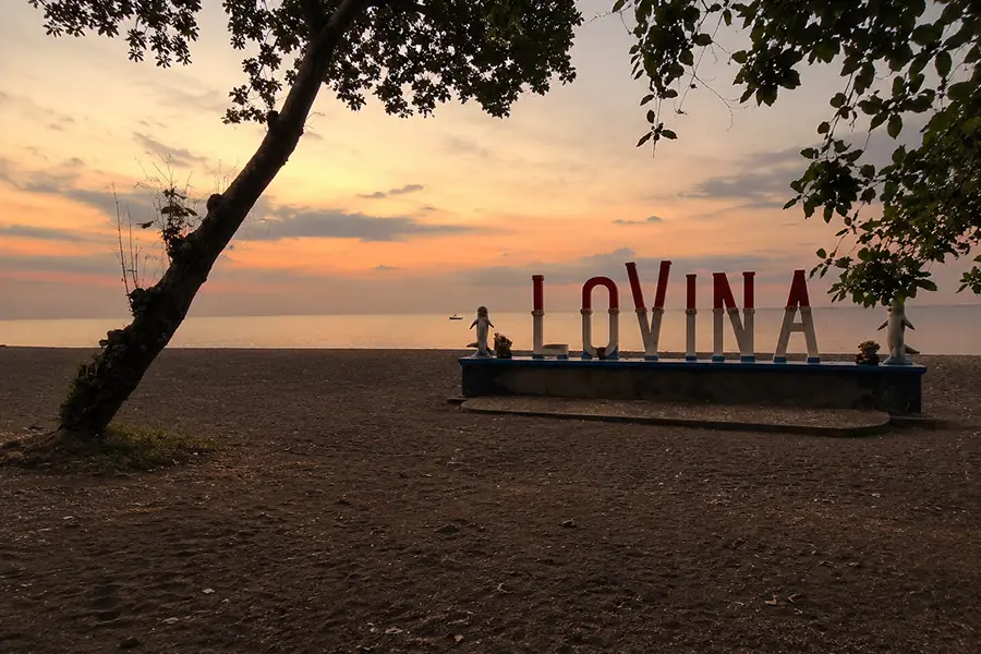 The iconic LOVINA sign landmark framed by leafy tree branches on the dark sand beach at sunset.
