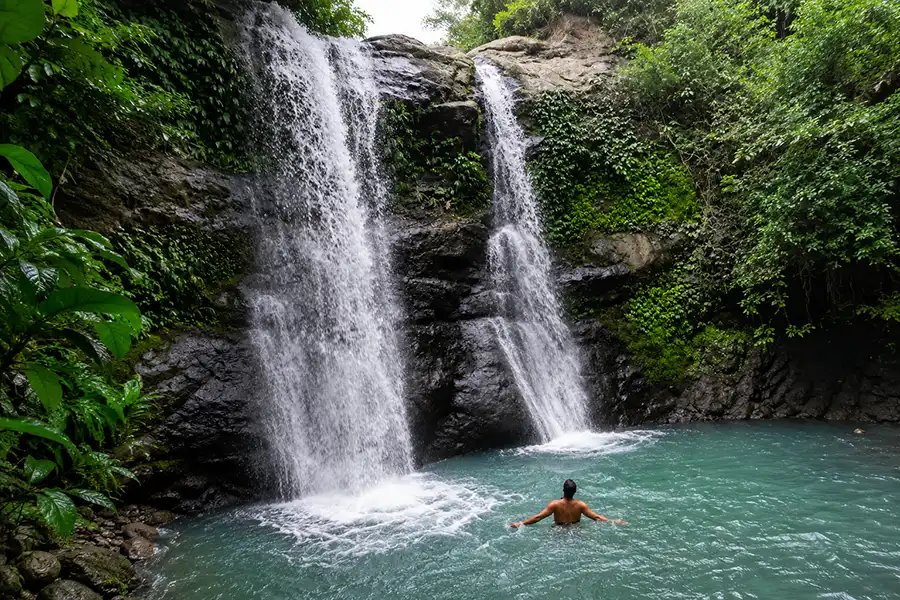 A swimmer with arms open enjoying the cool turquoise water in front of a twin cascade waterfall.