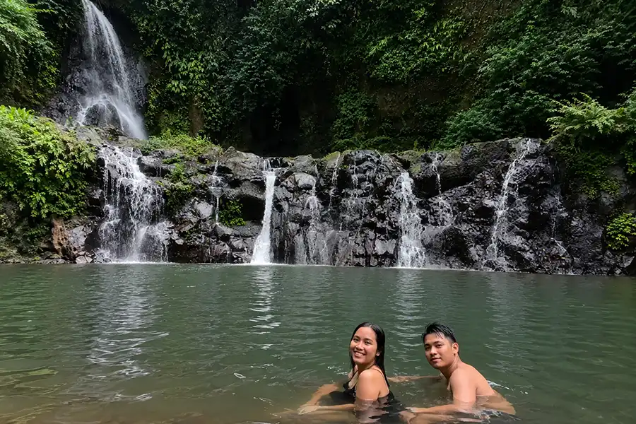 A couple enjoying a swim in a wide river pool with multi-tiered rock waterfalls in the background.