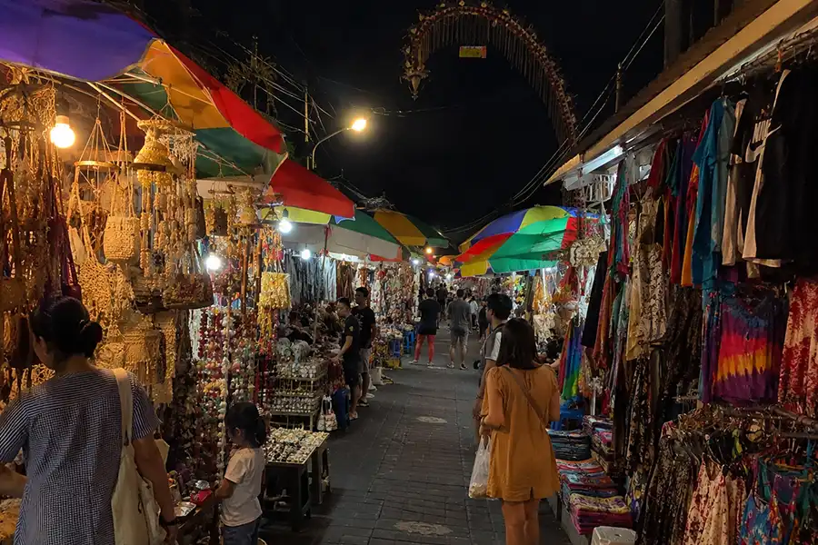 Tourists walking down a narrow street lined with colorful market stalls selling clothes and shell crafts.