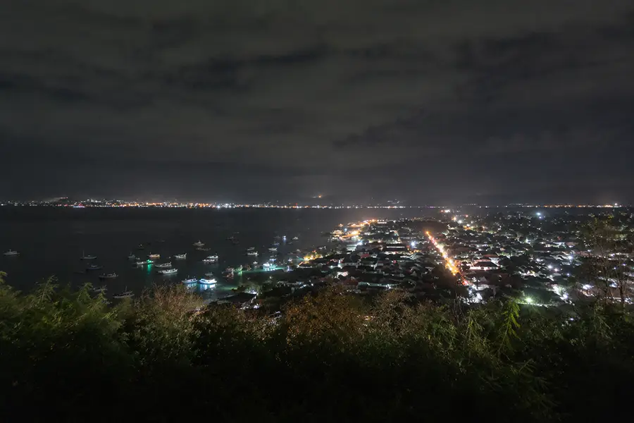 A night view from a high hill looking down at the glowing lights of a coastal town and boats anchored in the dark bay under a cloudy sky.