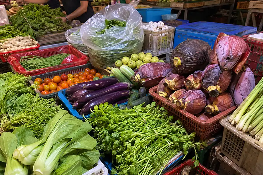A colorful array of fresh vegetables including eggplants, leafy greens, tomatoes, and banana flowers in market crates.
