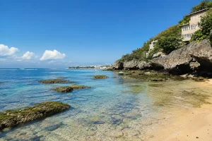 Bright yellow beach umbrellas shade wooden sunbeds along the sandy shore facing ocean waves. Lounging under the umbrellas at Padang Padang Beach Uluwatu is a popular way to unwind by the natural ocean shoreline.