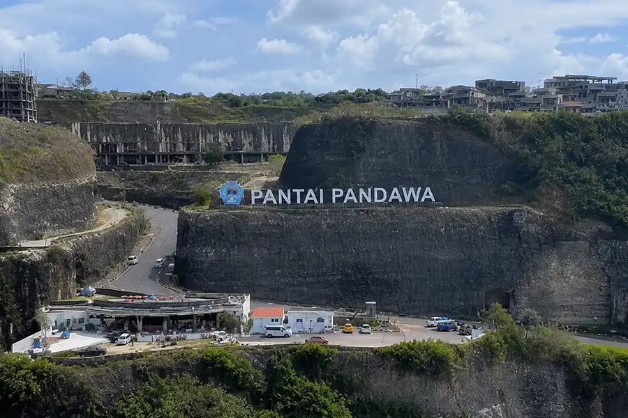High-angle view of huge limestone cliff cutouts featuring the large white PANTAI PANDAWA sign and winding access road.