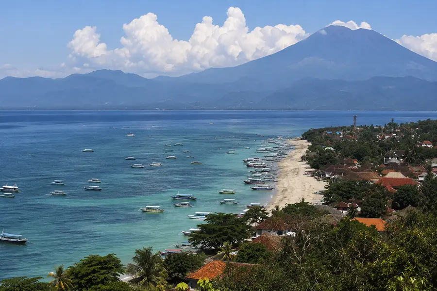 A high-angle view of a sandy beach lined with dozens of small boats, with a massive volcanic mountain rising in the background under a blue sky.