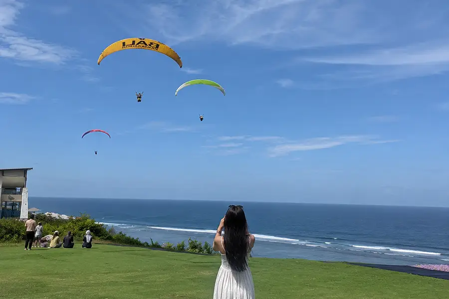 A woman taking a photo of paragliders soaring over the ocean from a grassy cliff.