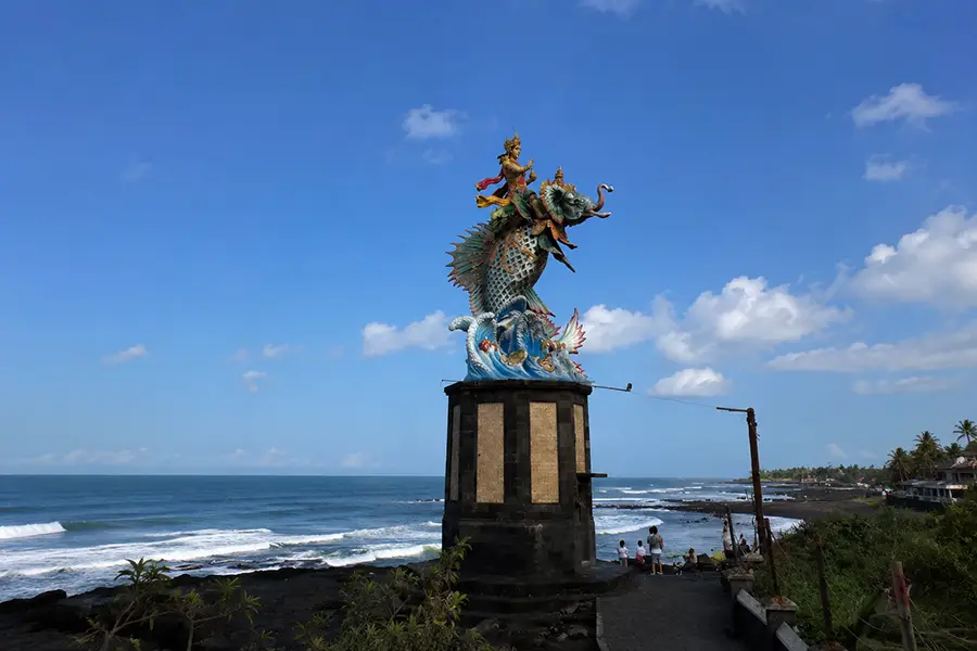 A tall mythological statue on a stone pillar overlooking a rocky shoreline and blue sea.