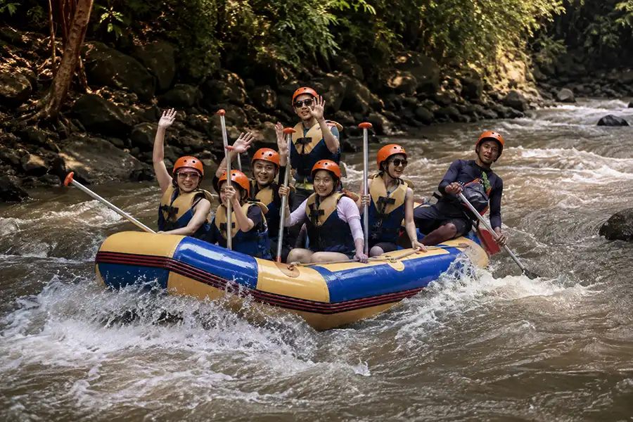 A group of tourists in orange helmets waving happily from a blue and yellow raft on a river.