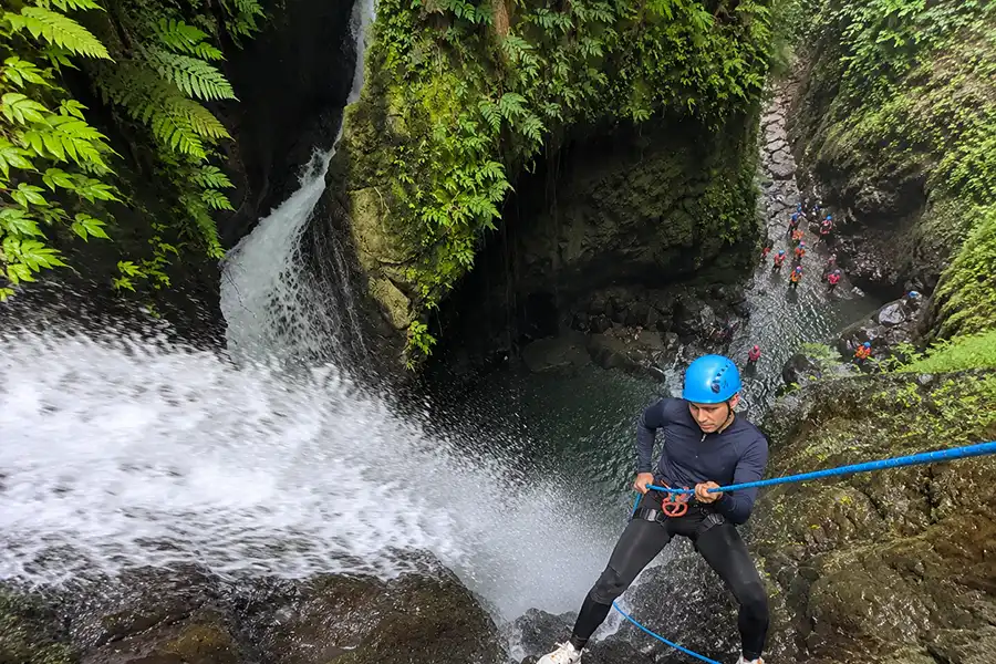 A male adventurer wearing a blue helmet rappelling down a steep cliff right beside a powerful gushing waterfall.