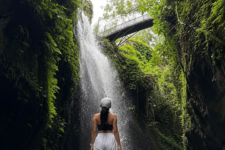 A woman in a white cap looking up at a tall waterfall and a metal pedestrian bridge above the gorge.