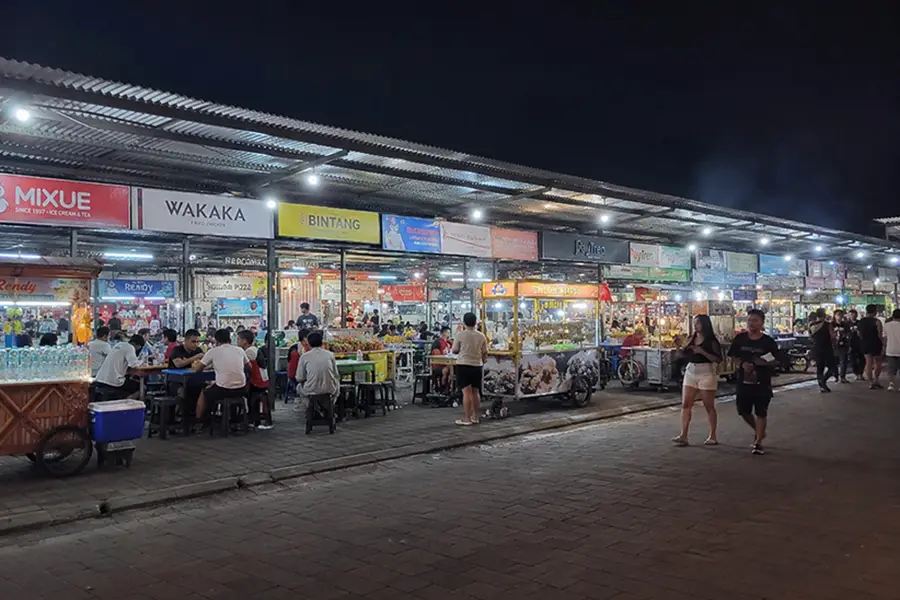 People walking and dining at open-air food carts under a long covered canopy at night.