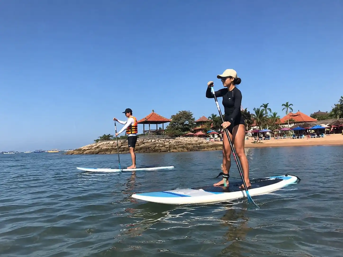 A woman and a man on separate stand-up paddleboards paddle in calm ocean water near a stone grotto on a clear day with traditional structures on shore.