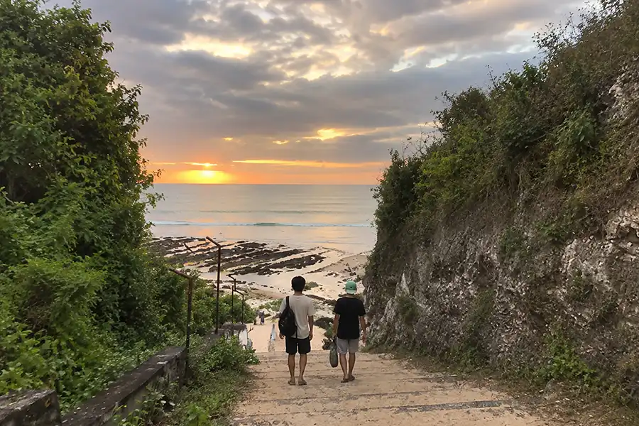 Two travelers walking down concrete stairs between green cliffs toward the ocean sunset.