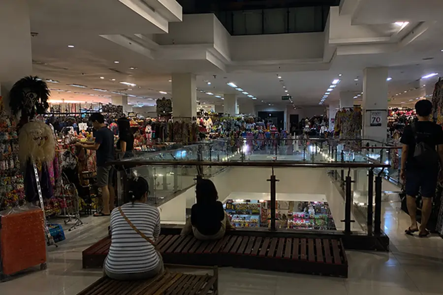 Shoppers browsing traditional clothing and crafts on the upper floor of a multi-level indoor market.